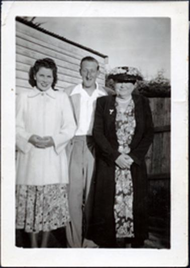 This image shows a man standing between two women, posing for a photograph. The man is wearing a light-coloured suit with an open-necked white shirt.  The younger woman is wearing a three-quarter length white coat over a floral dress.  The older woman is wearing a floral dress and a dark coloured full-length coat.  She is wearing a hat, necklace and has a brooch pinned to her coat.  They are all smiling.  Part of timber building is behind them as well as a wooden fence.  Tall shrubs can be seen behind the fence.  