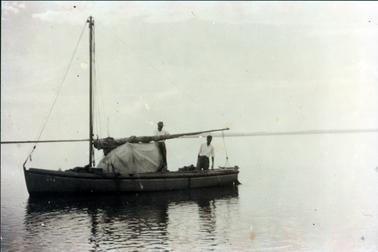 This image shows two men standing in a fishing boat on the water.  Land can be seen in the distance.  The boat has a tall mast.  The man standing on the left-hand side is wearing a white shirt, dark trousers and a hat.  He is standing in front of a covered hood behind a rolled-up sail.  The man on the right-hand side is wearing a white shirt and dark trousers.  There are ripples on the water along with the boats reflection in the foreground.  The image is not clear.  