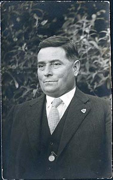 The image depicts a portrait of a middle aged man with dark hair and dressed in a suit with a fob watch and RSL badge on coat lapel.   