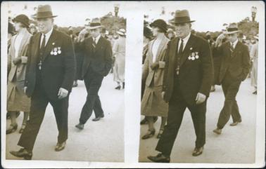 This photograph has two identical images, side by side.  The image shows a man in a three-piece suit walking along a street.  He is wearing a hat and smoking a cigarette.  He is wearing three medals on his coat and a badge in his coat lapel.  A fob watch is attached to his waistcoat.  Other people are walking along beside and behind him.   