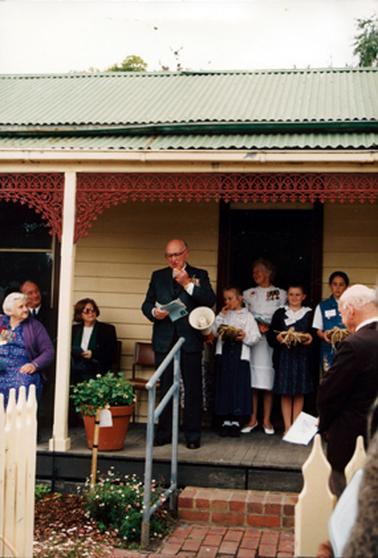 This image shows a group of people on the verandah of a wooden cottage.  The cottage is painted cream with a green iron roof.  The verandah has wooden floorboards, cream posts and brown fancy wrought iron trim.  A pipe railing is visible beside a brick step and path.  A man, dressed in a suit and wearing medals, is standing in the middle of the verandah reading from notes held in his right hand.  A megaphone is on his left-hand side.  Three people are sitting on the left-hand side of the verandah.  A woman, dressed in white with medals pinned on her dress, is standing in the doorway of the cottage.  Three young girls are standing in front of her.  The girls are holding baskets in their hands.  Part of a man, holding a white paper, can be seen on the right-hand side in the foreground.  