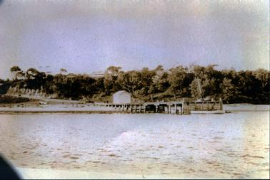 This image shows a wooden jetty.  The photo is taken from the water, looking back towards the land.  Timber planks are across the end of the pier and a boat is tied up beside them.  There appears to be a small crane on the end of the jetty.  Two people are walking from a motor vehicle towards a small shed at the land-end of the jetty.  A road runs down to the foreshore beside an embankment which is covered in trees.  Trees run down to the water on the right-hand side of the image.  The faint outline of a building can be seen in the far distance.  