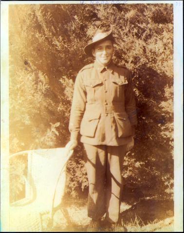 This image shows a young man dressed in army uniform standing on the grass in front of large shrub, posing for a photograph.  He is smiling for the camera and has his right hand resting on a white wicker garden chair.  