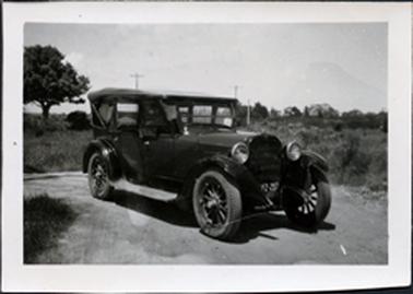 This image shows a four-door, soft-top car, parked on dirt. It has spokes in the wheels and  part of the number plate is YZ-297.  There is grass and trees in the background.  Two poles can also be seen behind the car.  