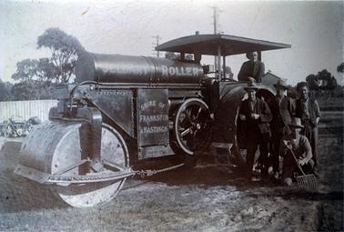 This image shows five men posing for a photo in front of a large steam roller.  One man is sitting on the machine, three men are standing beside the large back wheel and one man is crouching on the ground, holding a pitchfork.  All the men are wearing hats except for the man standing on the right-hand side who is wearing a cap.  The following words are printed across the side of the steam roller: “Jelbart Roller Shire of Frankston & Hastings”.  A motor cycle is standing beside a tall wooden fence on the far left-hand side.  Part of a small building can be seen behind the men and tall trees are in the background.  
