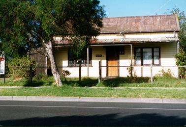 This image shows a weatherboard cottage with a faded red roof.  It has a four post verandah across the front. A set of three windows are either side of a wooden front door.  White curtains are at the windows on the right-hand side.  A plaque is on the wall on the left of the door and a electricity box is on the wall on the right-hand side of the door.  A high brown timber fence is along the left-hand side of the cottage.  A post and wire fence runs across in front of untidy grass.  A leaning tree is on the footpath and a bitumen street, with a white line, is in the foreground.  Part of a sign on the footpath, can be seen on the far left-hand side. 