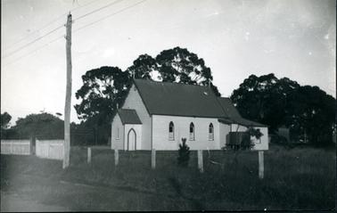 This image shows a wooden church with a galvanised iron roof and a cross on the top. There is a water tank on a stand on the side of the building.  The door into the narthex has a pointed top.  A white picket fence is seen at the front and continuing along from this is a post and wire fence in front of a paddock of grass.  A power pole is seen in the foreground and large trees are to the side and back of the church.  A tiny fur tree stands near the post and wire fence.  A small section of road can be seen in the foreground.  