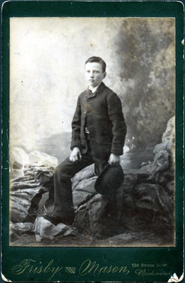 This image shows a teenage boy, dressed in a three piece suit, white shirt and tie, posing for a photograph.  He is holding a hat in his left hand and his right leg is bent with his right foot resting on a rock.  He is standing slightly side-on, on grass and there are rocks in the background.  He has a serious expression on his face.  Bushes can be seen on the right-hand side of the image.  The photographers details are stamped in gold writing at the bottom of the mounting board.  