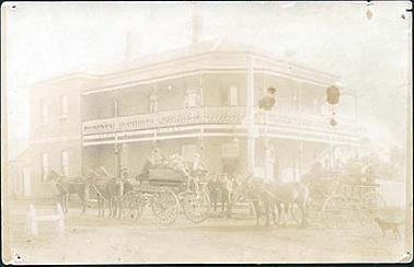 Royal Hotel, Hastings, built on both sides of a corner block. A large two story building with a verandah around both sides on both stories. The upper verandah has a decorative wrought iron strip around its lower edge. A back section has a plain facade with the word “billiards” on it. In front of the hotel are two open four wheeled carriages, one drawn by three horses, the other by two, each containing a group of men. Two men are standing next to the carriages, and three women are on the upper balcony. There is a dog in the right hand corner.