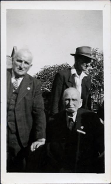 This image shows three men with trees in the background.  Two men are standing and one is sitting.  The two men at the front of the image are looking at the camera and are dressed in three piece suits, white shirts and ties.  They both have very little grey hair.  The man behind them is not looking at the camera and is wearing an open necked white shirt with the collar over his jacket.  He is wearing a hat.  