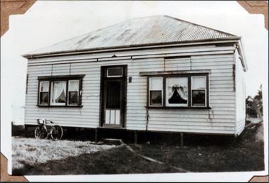 This image shows an old house taking up most the photograph.  It is of timber construction and has an iron roof.  A set of three windows are on either side of the front door.  There are blinds and lace curtains at the windows.  The house is on low stumps and a block of wood in used for a step at the front door.  A ladies bicycle with a basket on the front, is leaning against the house.  There is grass in the foreground and trees can be seen in the background on the far right-hand side of the photograph. Photograph ‘corners’ are seen  on each corner of the photograph.  
