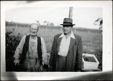 This image shows two men, from the hips up, standing side by side.  The man on the left of the photograph is elderly and wearing an open-necked shirt, overalls and a waistcoat.  He has a pipe tucked into  his belt and a walking stick in his right hand.  The other younger man is taller and is wearing an open-necked shirt, suit and hat.  There is a white container on the ground to his left.  There is a paddock of grass behind them and a fence and trees can be seen in the far background.  There is a shrub to the right of the elderly man and a post behind the other man.  