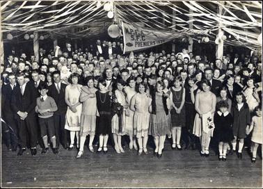 This image shows a very large group of people in a hall for a football celebration.  There is a large banner in the centre of the hall which reads: “M.P.F.A. S.F.C. Premiers 1928”.  Streamers and balloons hang from the roof.  Several wooden posts can be seen on either side of the hall and across the top.  The building is unlined and the galvanised iron roof can be seen.  The floor is wooden boards.  All the revellers are very well dressed - the men in suits and the girls in knee-length dresses with beads, white stockings and strappy shoes.   