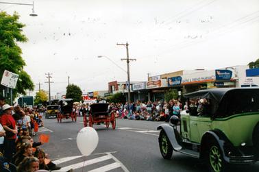 This image shows a parade of four vintage motor vehicles driving down the middle of a street, away from the camera.  People line both sides of the street.  Shop fronts can be seen on the right-hand side of the image including Bayne & Friend Pharmacy and the Hastings Bakery.  The vehicle at the front is yellow with a black roof, the next two vehicles are black with red wheels and the vehicle closest to the camera is green with a black roof.  A small white dog can be seen inside the green car.  A white balloon is in the foreground.  High electricity poles line the right-hand side of the street. 