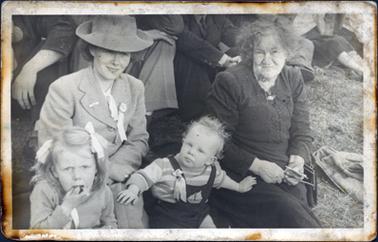 This image shows an elderly woman sitting on the grass with a young woman and two children.  The young woman is dressed in a suit and hat.  She has a bar brooch pinned to her right lapel.  The elderly woman is wearing a dark coloured dress with a brooch pinned at the neck and she has a handbag over her left arm.  She is holding something in her hands.  The little girl is eating.  She is wearing a jumper and has two bows in her hair.  The little boy has a striped jumper under his overalls which have a sailing boat at the front.  The young woman and the children all have a rosette and ribbons pinned on to their clothes.  The legs of other people sitting on the grass can be seen behind them.  
