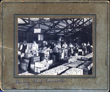 This image shows a packing shed with male and female workers packing boxes with apples. The workers are wearing aprons.  There is a man on the far left-hand side in a long sleeved white shirt, tie and vest.  All the boxes in the foreground are full.  There are many empty boxes beside the workers who are standing one behind the other.  More full boxes can be seen behind them.  The shed has a high sloped galvanised iron roof and all the timber beams are exposed.  Painted in white on one of the beams is: “May here first 1323-24”.  An ‘Important’ notice hands from another beam.  The photographers details are stamped on the bottom left-hand corner of the mounting board.  Written across the bottom of the mounting board in blue biro is: “Packing Shed Somerville”.