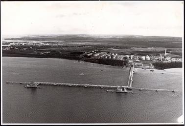 This image shows a town and bay taken from an aeroplane.  The long foreshore winds across the centre.  A pier and marina are at the centre of the image.  Several other boats are anchored in the bay.  Large tanks can be seen on land jutting out into the bay.  A large mangrove and park area is in the foreground on the left-hand side of the image.  