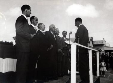 This image shows a group of six men standing before a seventh man at a swearing-in ceremony.  They are all holding a book in their right hand and the seventh man is reading from a sheet of paper.  Two microphones on stands are beside him.  Wooden buildings can be seen on the right-hand side of the photograph and people are standing in front of them, including a girl, holding a bouquet of flowers. 