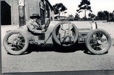 This image shows an elderly man sitting behind the wheel of a racing car looking at the camera.  He is wearing a light coloured coat and a battered hat.  He has a pipe in his mouth. The car has a spare wheel  attached to the side of the car and a rucksack is attached to the wheel with a leather strap.  A galvanized iron shed is behind the car on the left-hand side of the image.  Signs can be seen along the side wall of the shed including the word ‘shell’.  A small tank at the front of the shed also has the word ‘shell’ painted on it.  Two tall trees and other buildings can be seen the background and gravel is on the ground in the foreground.  