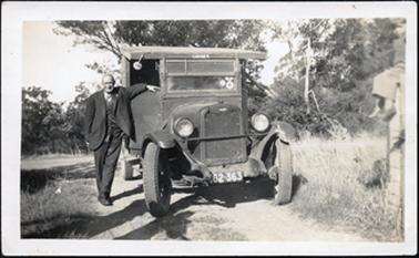This image is of a man posing beside a Chevrolet truck which is parked on a dirt road.  He is wearing a suit with a cardigan underneath his coat.  His arm is resting on the truck door.  A boy can be seen sitting on the top of a fence post at the far right of the photograph.  Trees can be seen in the background.  The number plate of the truck is 62-363 and the word ‘CARRIER’ can be seen above the windscreen.  