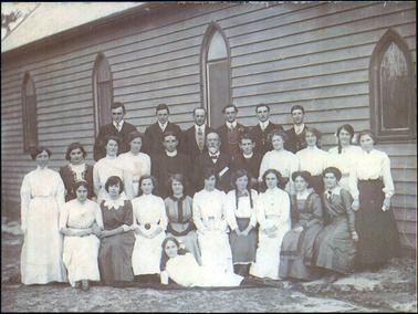 This image shows a group of twenty-seven men and women of all ages, posing for a photograph outside a church building.  There are six men standing on a form in the back row, eleven men and women standing in the middle row and nine women sitting in the front row.  A young woman is half lying on a rug at the front.  Everyone is beautifully dressed.  All the women have long dresses with long sleeves and high necklines and collars.  The men are all dressed in three piece suits with fob chains.  An older man in the middle row has a white beard and moustache.  The men, either side of him, are both ministers of the church.  The wall of the church is weatherboard with four church windows along its length.  