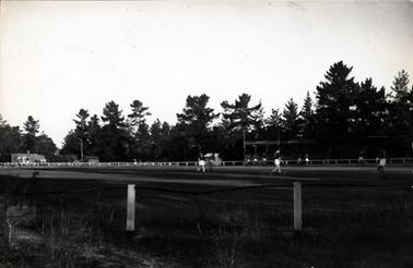 This image shows an overall photo of a football field with some players visible in the distance.  A spectators stand can be seen in the distance and large pine trees circle the oval.  Two posts with pipe between them can be seen in the foreground and a post and rail fence can be seen in the background surrounding the oval.  