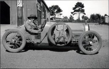 This image shows a man sitting in a racing car posing for the camera.  There is a large shed behind him on the left-hand side of the image.  A building can be seen behind a line of trees, both big and small, in the background behind a fence.  Another small building can be seen on the right-hand side of the image.  A petrol bowser is at the front of the shed and the word SHELL can be seen along the side of the shed.  