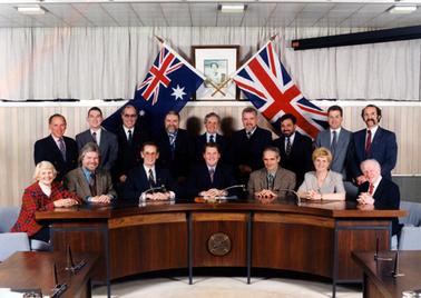 This image shows a group of fourteen men and two women sitting and standing behind a desk posing for a photo.  The curved desk is polished timber and has a round plaque on the front.  Everyone is smiling for the camera.  Nine of the men are standing.  Everyone is well dressed.  All of the men are wearing suits and ties.  Two flags are crossed behind the group in front of a photo of the Queen.  Curtains are across a set of windows in the background above a timber panelled wall.
