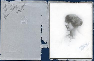 This image is of a portrait of a young woman taken from a side angle from the chest up. She  is wearing a white square necked top and has a fine chain around her neck.  Her hair is swept up and pinned at the back.  The photographers details are written across the bottom left-hand side of the photograph.  The photograph is mounted on board and glued into a brown paper cover.  A royal crown is embossed on the front of the cover and the photographers details are printed in brown ink .  