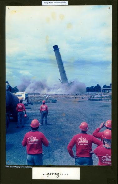 This image shows a tall chimney stack leaning to the left after blasting.  Thick grey smoke is swirling around its base.  Six workmen are standing watching with their backs to the camera.  They are all wearing blue pants, red long sleeved shirts and red helmets.  The words “Able Blasting The Master Blaster” is printed on the back of their shirts.  Part of a building can be seen on the far right-hand side.  A white vehicle is on the left-hand side along with a tank on a trailer.  A man is sitting on the top of the tank taking a photo.  