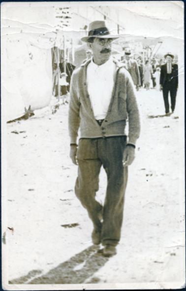 This is an image of a man walking along outside a marquee.  He is looking away from the camera and is mid-stride.  He is wearing a white shirt, done up to the collar, cardigan and dark trousers and a hat with a very wide band.  He has a large moustache and is wearing spectacles.  There are many people in the background, including two men wearing suits, ties and hats who are walking a short distance behind him.  
