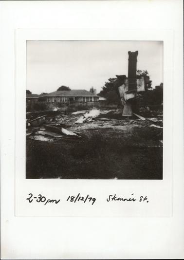 This image shows the burnt-out remains of a building.  A brick chimney is still standing and there is a lot of twisted sheets of corrugated iron around the chimney and on the ground.  A brick house with a tiled roof is behind a fence in the background.  Part of a roof of another house can be seen on the left-hand side of the image.  Tall trees can be seen in the background behind the chimney and there is grass in the foreground.  
