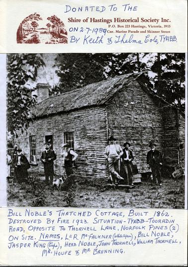 This image shows a group of men standing outside a wattle and daub cottage with a thatched roof, posing for a photo.  There are four men and a dog on the left-hand side of the cottage and three men on the right-hand side.  There is man standing in the doorway and a set of windows on either side.  A large chimney is at the back of the cottage.  Tall pine trees are behind the cottage and part of a tree can be seen in the foreground on the left-hand side.  