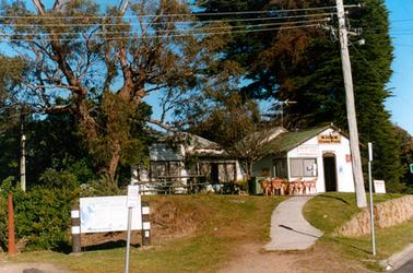 This image shows two buildings standing side by side beneath large trees.  The building on the right has the sign “Kiosk Stony Point” above the open doorway.  Other signs are on the front of the building.  The building on the left has windows right across the front.  A picnic bench table and stools are on the left and a table surrounded by plastic chairs is on the right.  A concrete path runs from the footpath to the front door of the Kiosk.  A large signboard is on the footpath to the left of the image and a power pole is on the right.  A street sign is on the footpath on the far right-hand side and a stone wall runs along the footpath behind it.  