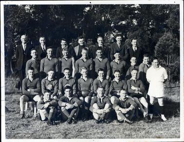 This image shows a football team posing for a photograph.  There are seven men standing on a form in the back row, eight men in the middle row, seven men sitting on chairs and four men sitting cross-legged on the grass.  A man in white is standing on the far right-hand side of the players.  Most of the men in the back row are wearing  white shirts, ties and suits.  There are tall trees behind the group.  One player in the centre of the photo is holding a football on his lap.