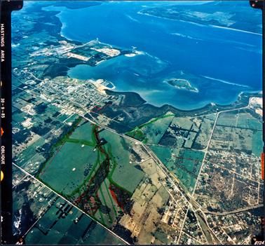 This image shows an aerial photograph of a town, foreshore and bay.  Streets, houses and paddocks can be seen.  A small island is in the bay and land can be seen in the top right-hand corner of the image.  Printed in white on a black border on the left-hand side of the image is: “Hastings area 30-9-85 Oblique”.  Areas have been marked out in red and green biro.
