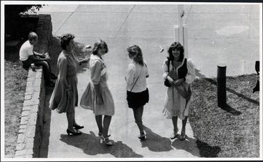 This image shows four women walking along a concrete path, half turning to look back.  Three of the woman are young and are wearing a sash.  The other woman, on the left of the girls is older.  A boy is sitting on a brick retaining wall looking at them.  A white pole is behind the girl on the right.  