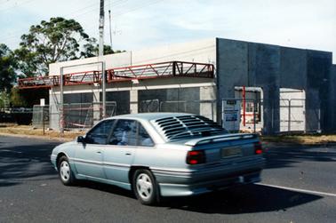 This image shows a construction site and a partly built concrete building. The top of a tall crane can be seen behind the building and a bitumen street is in the foreground.  Power lines are across the top of the building and trees can be seen on the left-hand side of the image.  Shadows from trees are across the street.  A silver coloured sedan motor car is driving along the street.  Windswept clouds are in the top half of the image.  