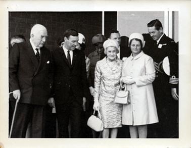 This image shows a group of men and women standing.  There are two men and two women at the front and part of six people can be seen standing behind them. The two men at the front are dressed in dark coloured suits, white shirts and ties.  The man on the far left-hand side is holding a walking stick in his right hand.  The woman, second from the right, is wearing a two piece print suit, hat, gloves and pearls and is holding a handbag by her side.  The other woman is wearing a coat and hat and has a handbag over her right wrist.  Her hands are clasped at the front.  A man in uniform, with his hat under his arm, is on the far right-hand side of the image.  