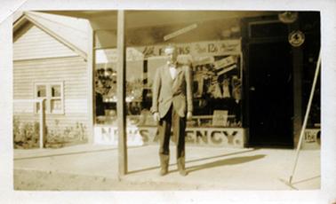 This image shows a man standing outside a shop, posing for a photo.  He is wearing dark trousers, a lighter coloured coat, white shirt and a bow tie.  A large front window and the front door of the shop can be seen.  The shop window is full of goods and signage.  ‘Newsagency’ is printed in very large letters across the bottom of the window.  More advertizing is across the top of the window including the words “Frocks”.  A verandah post is to the right of the man and a broom is standing to the left.  Part of a wooden building with a square window at the front is on the left-hand side of the image.  A garden and post and rail fence is  in front of the building.  