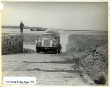 This image shows a cement mixer backed down an incline into water.  A stone retaining wall is on the left with a man standing on the top.  A bank of soil is on the right with a large boulder in the foreground.  Boats can be seen in the water and land in the background.  The number plate on the truck is JEE 135.  A piece of white paper with ‘Constructing the Boat Ramp - 1972’ is pasted to the bottom of the photograph.  Three more men can be partly seen around the truck.  