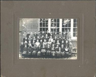 This image shows four rows of school children posing for a photograph.  There are twelve boys in the back row, eleven boys in the second back row, thirteen girls in the second front row and ten girls sitting in the front row.  One girl is holding a backboard which reads: “Hastings School No 1098 12-9-24”.  A brick wall with four large windows is behind them.  An air vent can be seen on the far left-hand side of the photograph.  Parts of wooden school forms can be seen on the right-hand side.