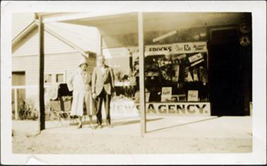 This image shows a man and woman standing outside a shop, posing for a photo.  The woman is wearing a full length light coloured coat and hat.  There is a pram behind her.  The man is wearing dark trousers, a lighter coloured coat, white shirt and a bow tie.  A large front window and the front door of the shop can be seen.  The shop window is full of goods and signage.  ‘Newsagency’ is printed in very large letters across the bottom of the window.  More advertizing is across the top of the window including the words “Frocks”.  Two verandah posts can be seen in the foreground.  Part of a wooden building with a square window and doorway at the front, is on the left-hand side of the image.  A garden and post and rail fence is in front of the building.  