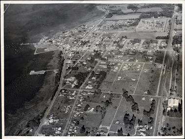 This image shows an aerial photo of a town with a shoreline and water on the left-hand side of the image.  A jetty can be seen with boats dotted on the water. Paddocks growing crops can be seen at the top of the image.  