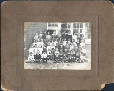 This image shows a group of school children, standing and sitting in five rows, in front of a building, posing for a photo.  There are eleven girls standing in the back row, thirteen girls standing in the second back row, thirteen boys and girls standing in the third back row, seven boys sitting on a form in the second front row and eight boys sitting, cross-legged on the grass, in the front row.  The small blackboard on the ground at the front reads: “S.S. 1098 (1) Hastings 17-2-21”.  The brick wall of the school with four large windows, is behind the children.   