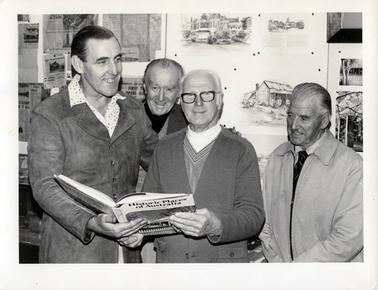 This image shows four men posing for a photograph.  There is a tall man on the left-hand side of the photograph, smiling and holding a book, “Historic Places of Australia”.  He is wearing a patterned shirt with the collar over the top of his buttoned-up jacket.  He has his hand on the shoulder of a man in the centre of the image, who is also holding the book.  This man is wearing a T-shirt, pullover and cardigan and wearing spectacles. There is a man standing behind and between them smiling broadly at the camera.  Another man on the right-hand side of the photograph is looking on and smiling.  He is wearing a shirt, tie and a jacket zipped at the front.  The wall behind the group is covered in pictures, paintings and newspapers articles.  