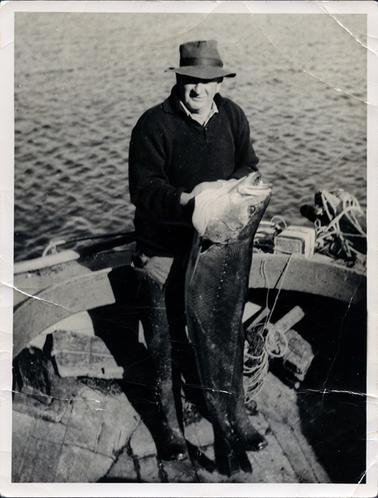 This image shows a fisherman standing in a wooden boat holding a very large fish and smiling for the camera.  He is wearing a dark coloured pullover and long rubber boots up to his thighs.  He has a hat pulled down over his eyes.  The fish stretches from his chest to the floor of the boat.  Ropes can be seen in the boat and the water is behind him.  