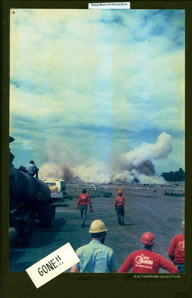 This image shows thick grey smoke swirling across the centre of the photograph after an explosion.  Five workmen are walking towards the site.  They are wearing, or carrying helmets and all except one man is wearing blue trousers, and red shirts with printing on the back that reads: “Able Blasting The Master Blaster”. The other man is wearing a short sleeved blue shirt and white helmet.  Another man, on the left-hand side, is sitting on the top of a tank taking a photo.  Part of two vehicles can be seen on the left.  