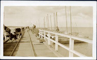 This image shows a jetty jutting out into the water.  The photograph is taken in the middle of the jetty looking all the way to the end.  A white post and rail fence is either side of the wooden jetty and rail tracks run down the middle.  Tied up to the jetty on the right-hand side is a row of approximately thirteen fishing boats with tall masts.  A man can be seen standing on one of the boats holding onto a mast.  People can be seen fishing off the jetty on both sides, along with dogs and fishing nets.  A wooden box is on the jetty in the foreground.  