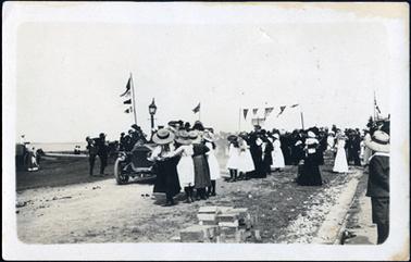 This image shows a large group of men, women and children, mostly with their backs to the camera, watching a car passing by.  Everyone is well dressed and wearing hats.  There are flags and banners flying.  A gas lamp can be seen behind the car and a horse and carriage is partly visible on the left hand side of the photograph.  Water can be seen in the far background.  There is a pile of bricks in the foreground.  
